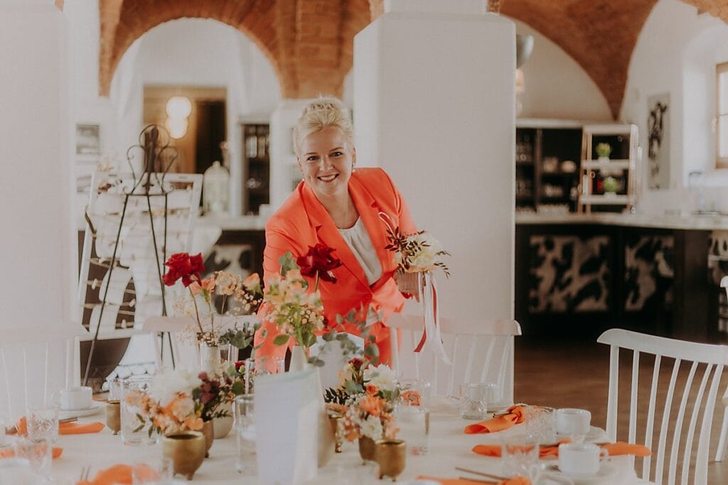 Eine lächelnde Frau in einem leuchtend orangefarbenen Blazer steht an einem mit Blumen geschmückten Esstisch, der für eine Veranstaltung in einem gemütlichen, eleganten Raum mit gewölbten Ziegeldecken gedeckt ist.
