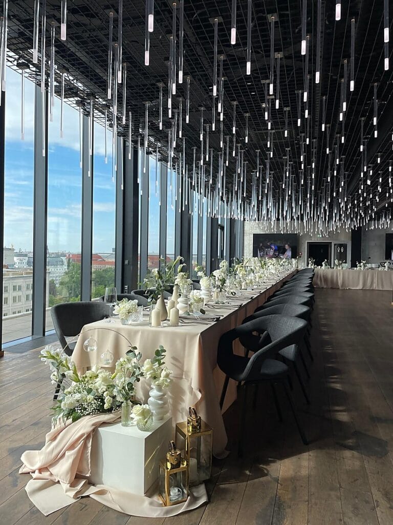 Eleganter Bankettsaal mit langen Tischen, die mit weißen Blumen, Grünzeug, Kerzen und beigen Tischtüchern gedeckt sind. Moderne schwarze Stühle säumen die Tische unter einer Decke mit Hängelampen, und große Fenster bieten einen Blick auf die Stadt.