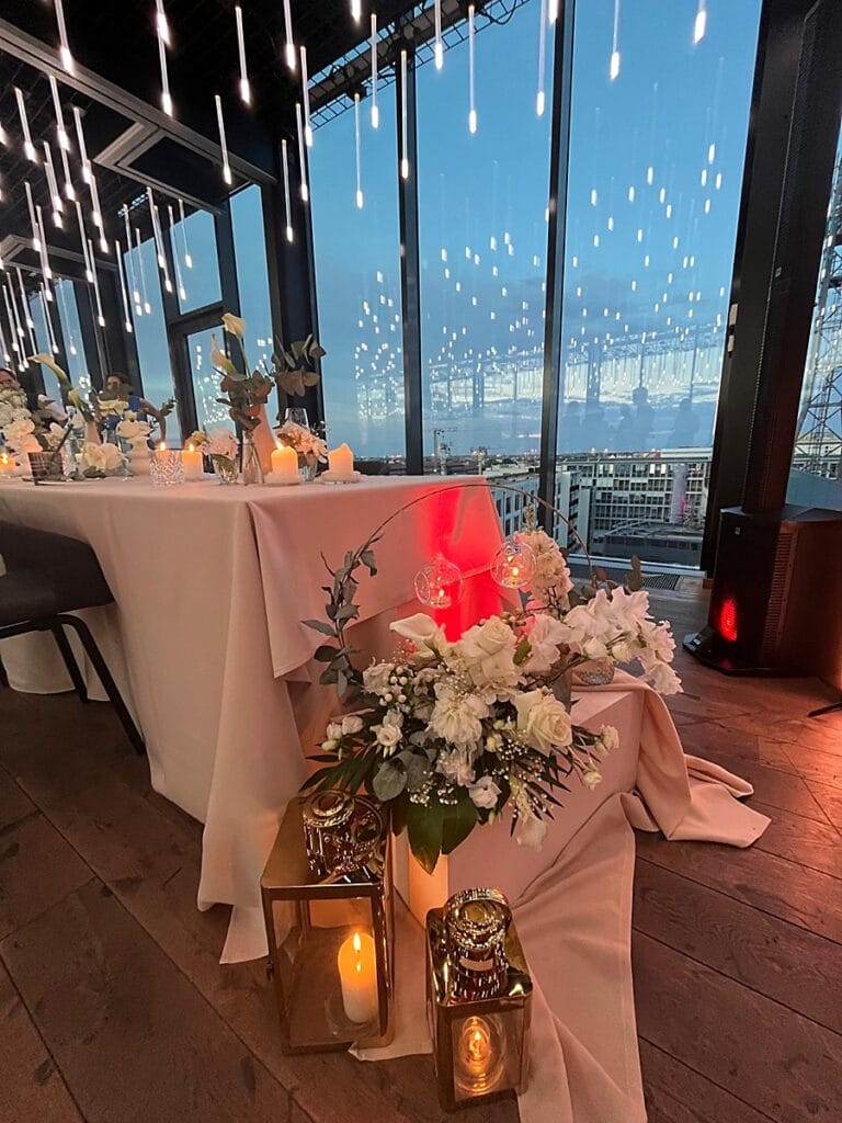 Eine dekorierte Hochzeitstafel im Innenbereich mit weißen Blumen, Kerzen, goldenen Laternen und einer erröteten Tischdecke vor bodentiefen Fenstern mit Blick auf eine Stadtlandschaft in der Abenddämmerung und Lichterketten an der Decke.