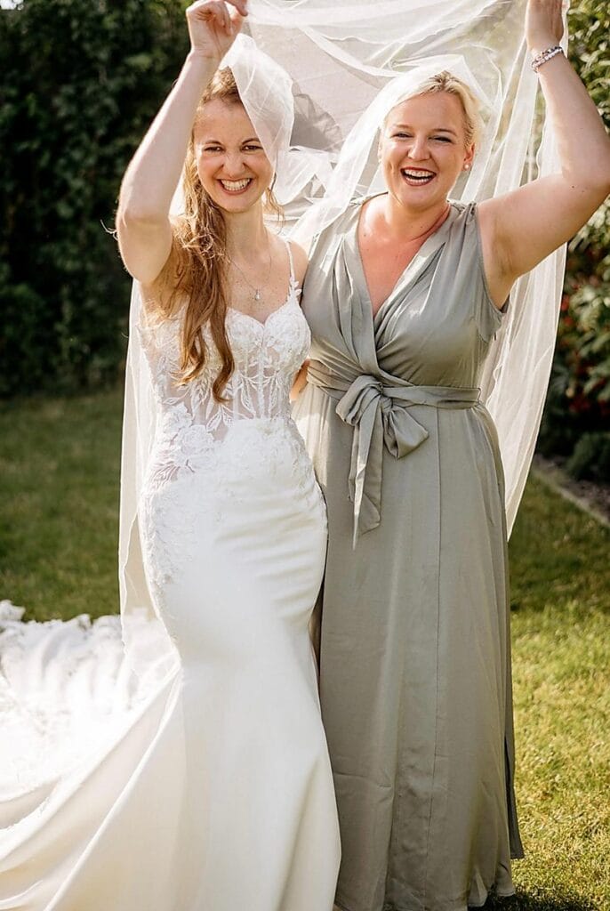 Zwei Frauen lächeln freudig im Freien; die eine trägt ein weißes Hochzeitskleid und einen Schleier, die andere ein hellgraues Kleid. Sie halten den Schleier gemeinsam hoch und stehen auf einer grünen Wiese im Hintergrund.