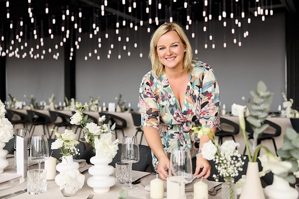 Eine Frau in einem geblümten Kleid lächelt, während sie einen mit weißen Blumen, Kerzen und Gläsern dekorierten Tisch unter Reihen von Hängelampen in einem modernen Veranstaltungsraum deckt.