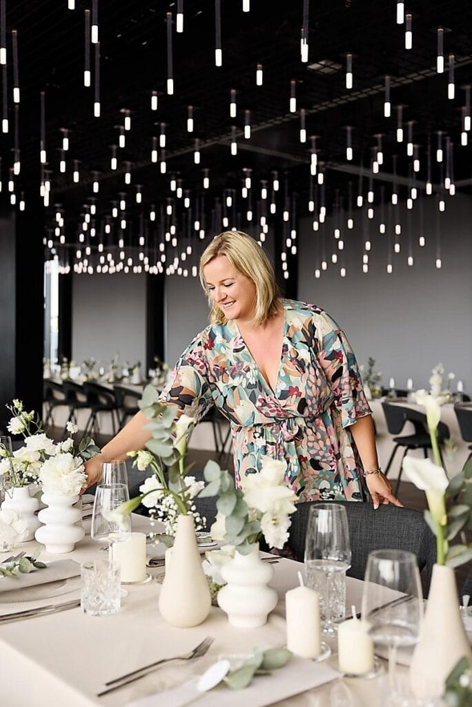 Eine Frau in einem Blumenkleid arrangiert Blumen auf einem elegant gedeckten Esstisch unter einer Decke aus Hängelampen in einem modernen, stilvollen Lokal.