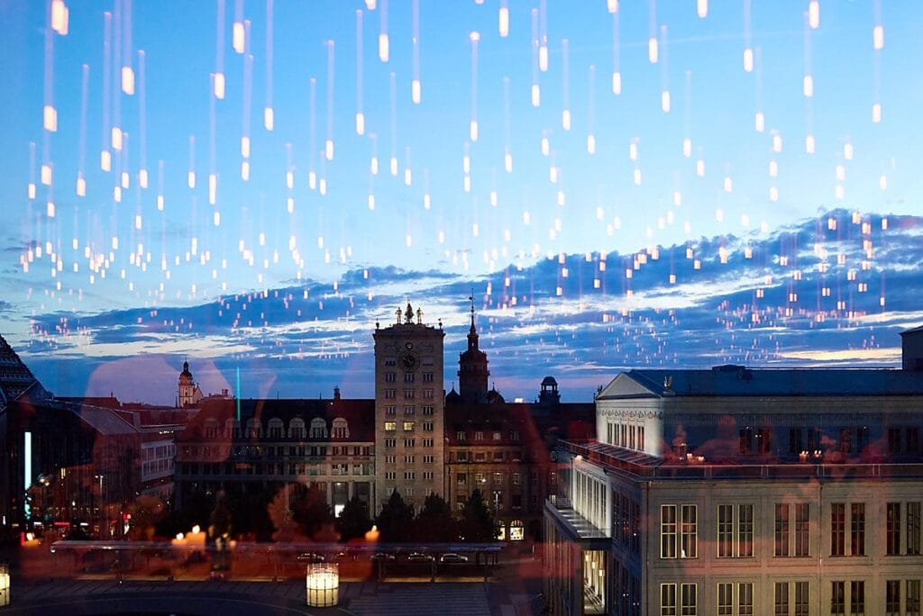Ein Stadtbild in der Abenddämmerung mit historischen Gebäuden unter blauem Himmel. Auf einem Glasfenster sind die Reflexionen vieler Hängelampen zu sehen, die eine Überlagerung von leuchtenden Punkten über der Stadt erzeugen.