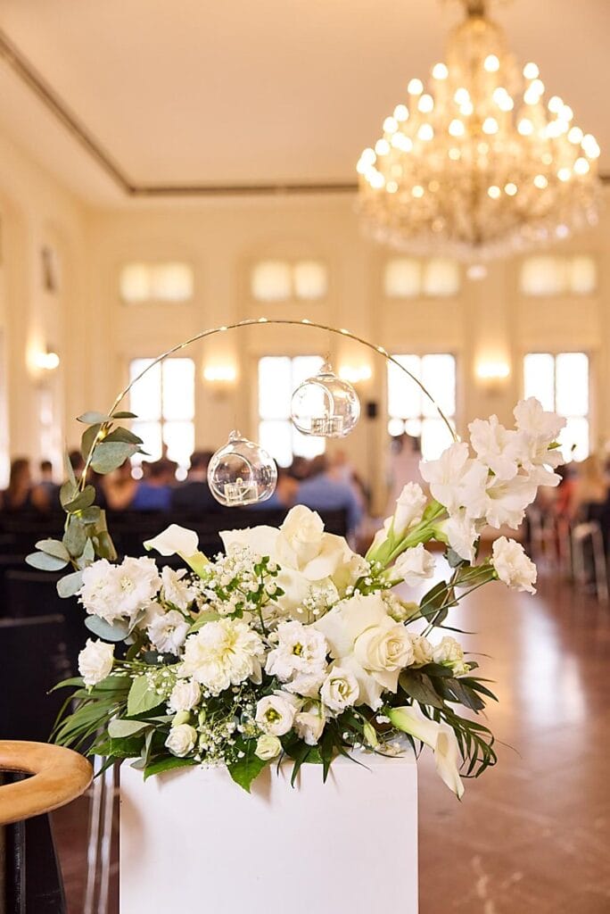 Ein weißes Blumenarrangement mit Rosen und Grünzeug steht auf einem Podest. Darüber hängen zwei Glaskugeln, und von der Decke hängt ein funkelnder Kronleuchter in einem sonnenbeschienenen, eleganten Raum mit sitzenden Gästen im Hintergrund.