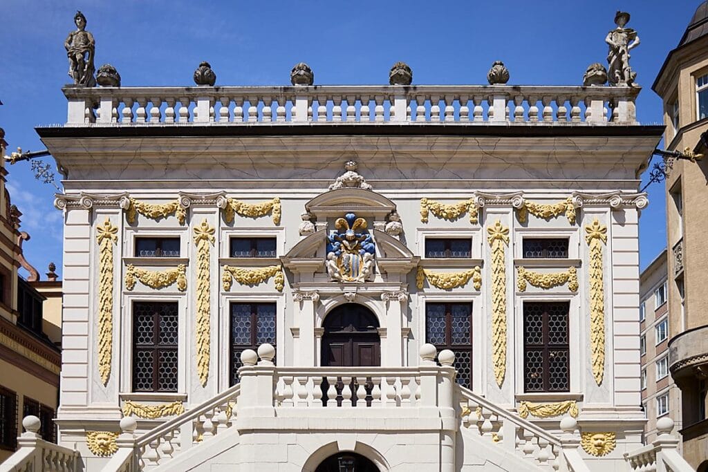 Ein Gebäude im Barockstil mit verschnörkelter gelb-weißer Fassade, dekorativen Statuen, Bogenfenstern und einer großen Treppe, die zu einem zentralen Portal unter einem geschnitzten Balkon führt.