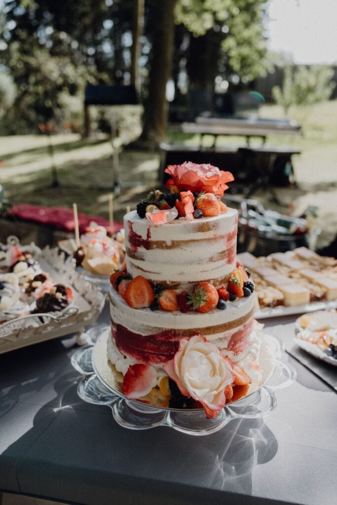Eine zweistöckige nackte Torte, dekoriert mit frischen Erdbeeren, Brombeeren und Blumen, steht auf einem gläsernen Kuchenständer im Freien, umgeben von verschiedenen Desserts und Grünzeug im Hintergrund.
