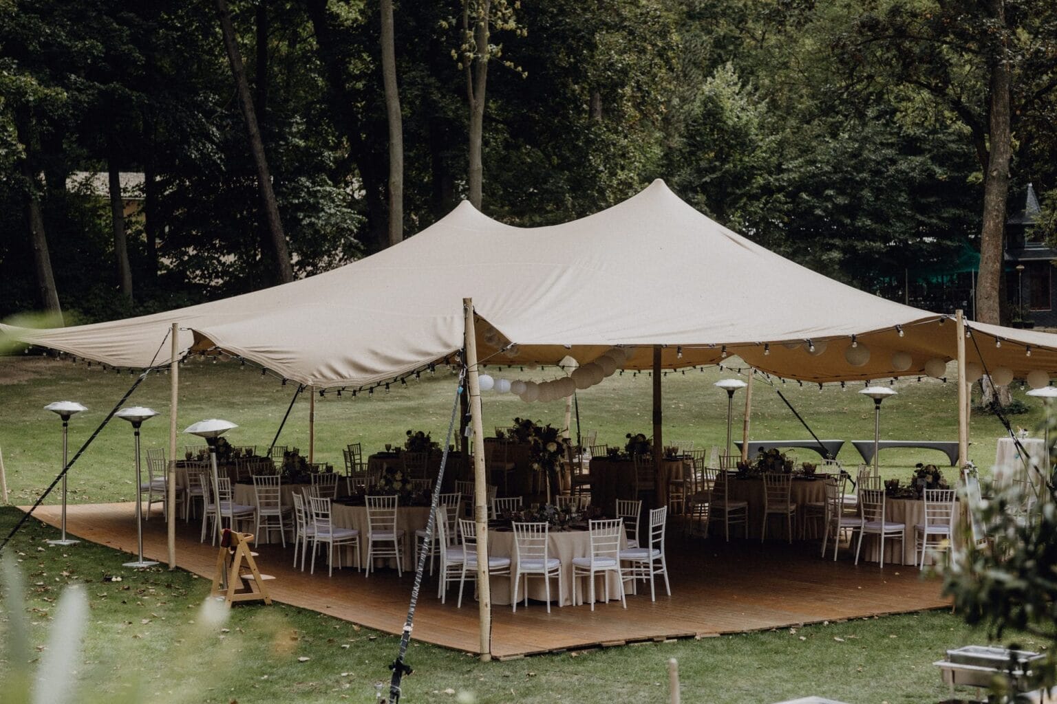 Ein großes, beigefarbenes Zelt ist auf einer Wiese aufgebaut und bedeckt runde Tische mit weißen Stühlen, die für eine Veranstaltung aufgestellt sind. Das Zelt ist von Bäumen umgeben und an den Rändern hängen Lichterketten.