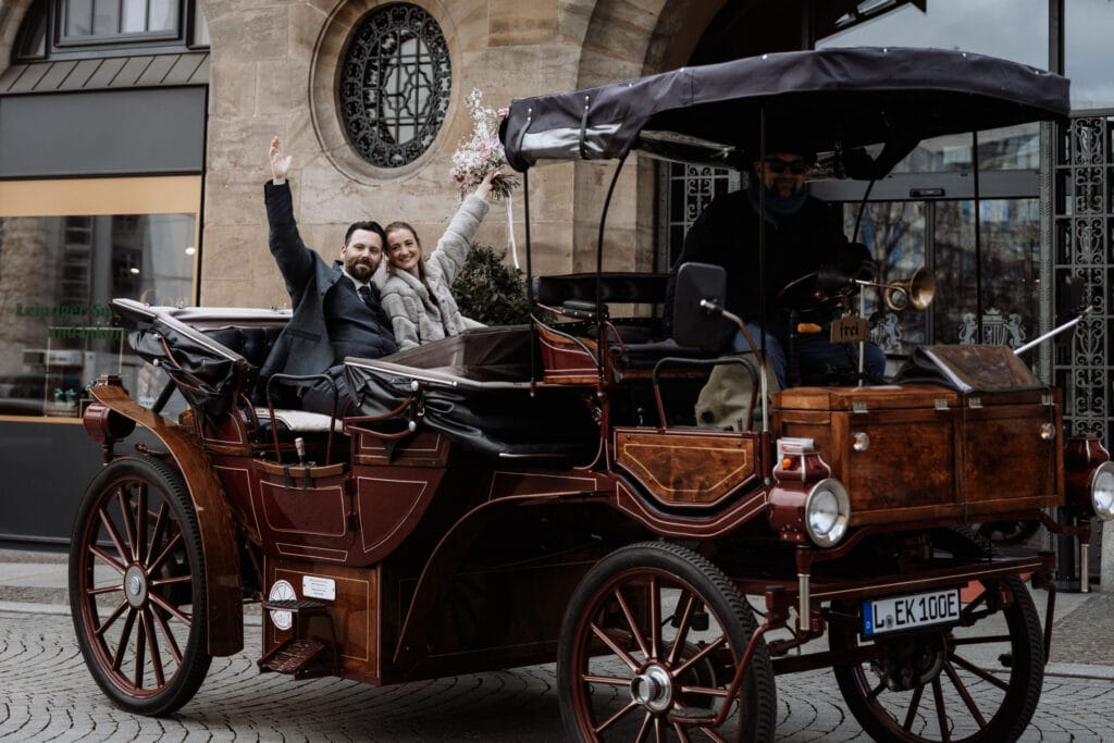 Ein Paar lächelt und winkt, während es auf dem Rücksitz eines braunen Oldtimerwagens mit Fahrer sitzt, der auf einer Kopfsteinpflasterstraße vor einem steinernen Gebäude geparkt ist.
